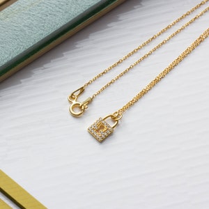 May include: A gold-toned chain necklace featuring a small, square-shaped padlock pendant. The padlock is embellished with tiny, clear, sparkling stones and has a central keyhole cutout. The necklace includes a circular spring ring clasp.
