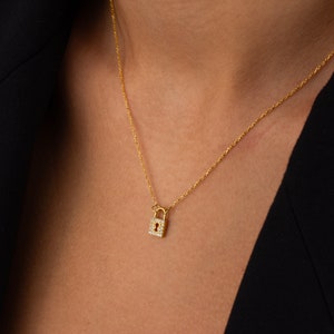 May include: A gold-toned necklace featuring a small, rectangular padlock pendant. The padlock is embellished with tiny, clear, sparkling stones along its border and displays a central keyhole cutout. The pendant hangs from a delicate gold-toned chain.