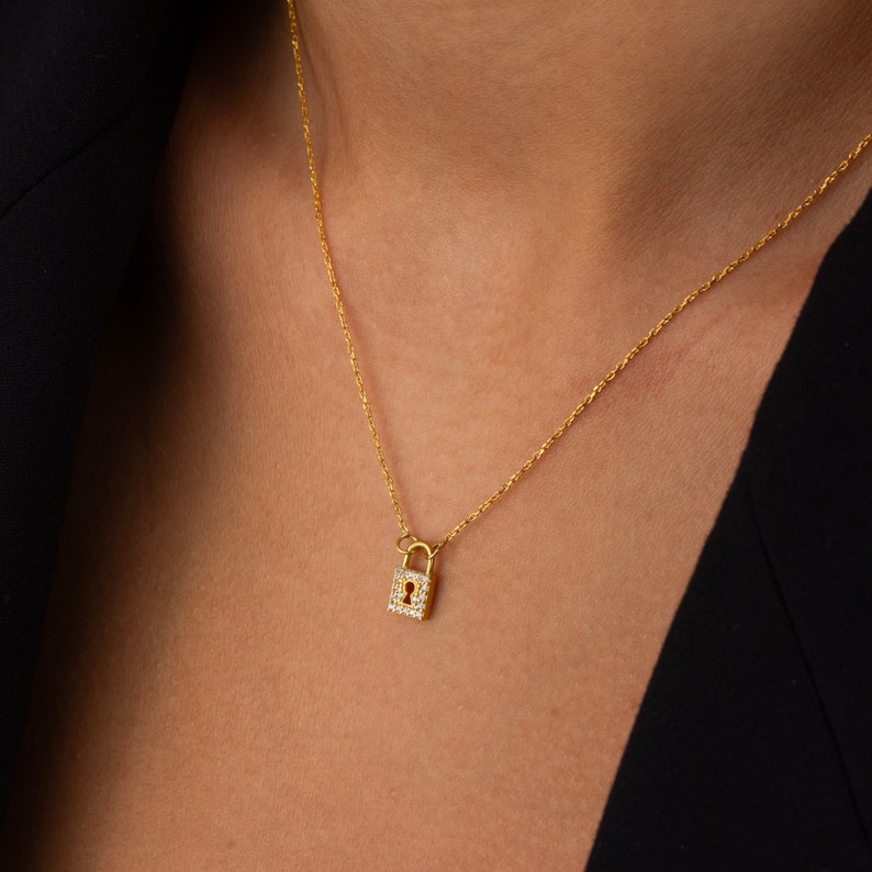 May include: A gold-toned necklace featuring a small, rectangular padlock pendant. The padlock is embellished with tiny, clear, sparkling stones along its border and displays a central keyhole cutout. The pendant hangs from a delicate gold-toned chain.