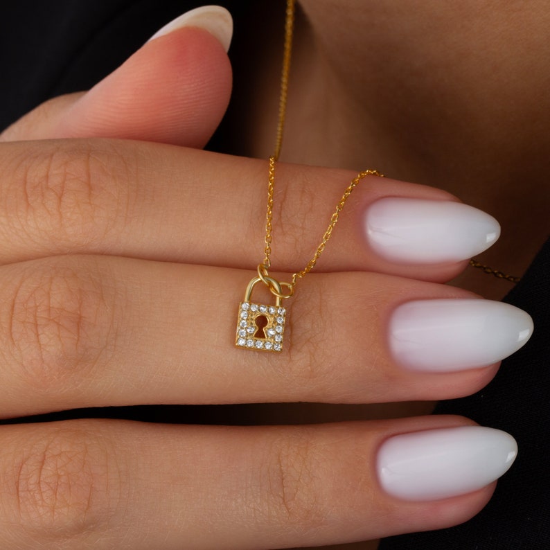 May include: A gold-toned necklace featuring a small, rectangular padlock pendant. The padlock is encrusted with numerous tiny, clear, sparkling stones and displays a central keyhole cutout. The pendant hangs from a delicate, thin gold-toned chain.