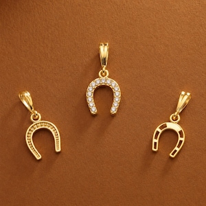 May include: Three gold-toned horseshoe pendants arranged in a triangular formation. The central pendant is embellished with a row of small, clear, sparkling stones along its arch, while the two flanking pendants feature a plain, polished gold finish.