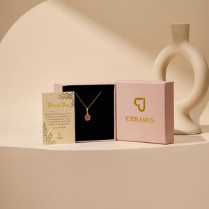 May include: A gold necklace with a small circular pendant featuring a red stone, presented in a light pink box with the gold text CERASUS and a logo. A small cream-colored thank you card with green leaf illustrations is placed next to the box.