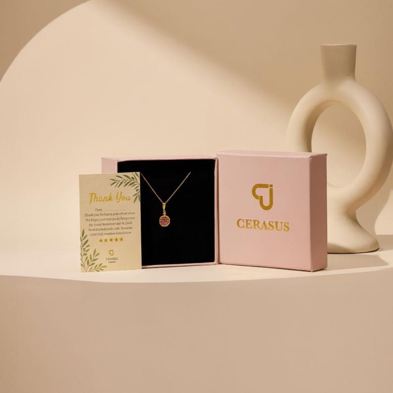 May include: A gold necklace with a small circular pendant featuring a red stone, presented in a light pink box with the gold text CERASUS and a logo. A small cream-colored thank you card with green leaf illustrations is placed next to the box.