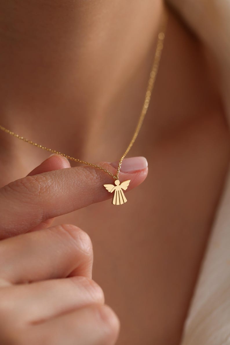 May include: A gold-toned necklace featuring a small, flat, stylized angel pendant with outstretched wings and a flared dress design. The pendant is suspended from a delicate, thin gold-toned chain that rests against the skin.