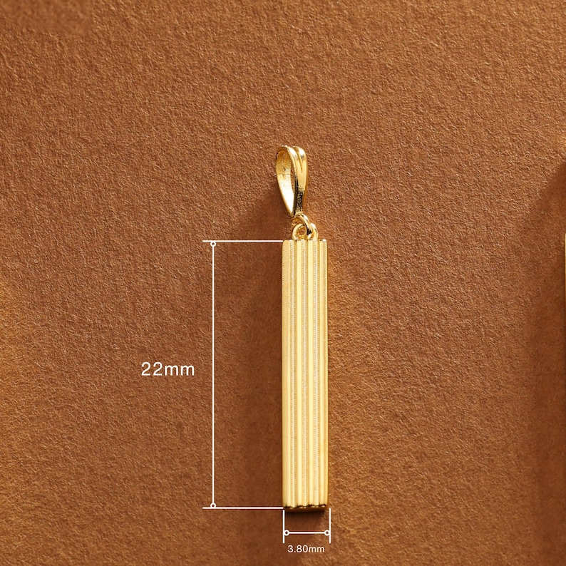 May include: A gold-colored pendant with a rectangular, vertically-lined design. The pendant measures 22mm in length and 3.80mm in width. It has a small loop at the top for a chain.