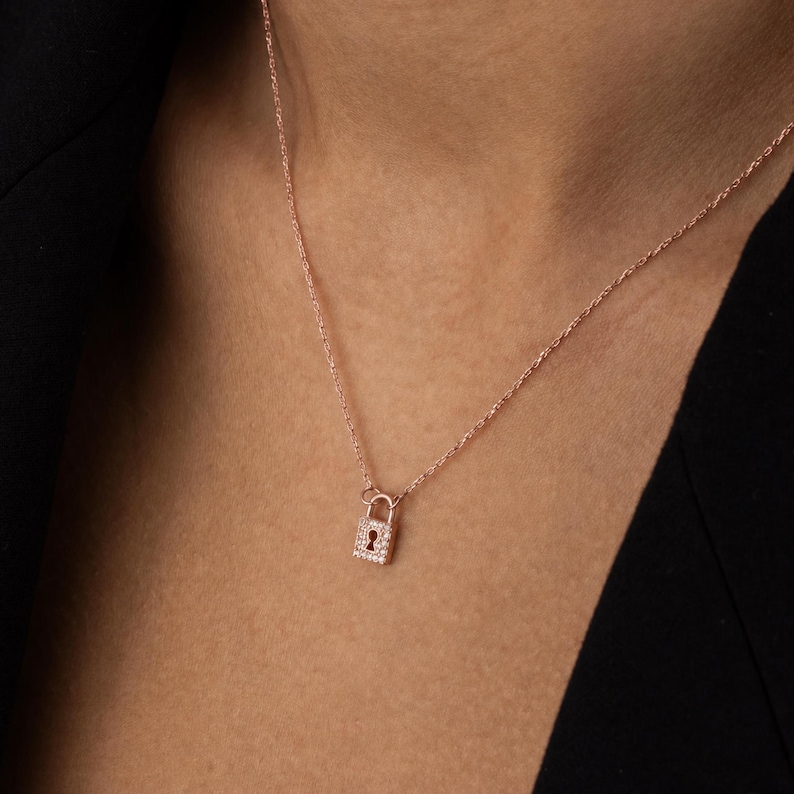 May include: A rose gold necklace featuring a small, rectangular padlock pendant. The padlock is encrusted with tiny, clear pav&eacute;-set stones and displays a central keyhole cutout. The pendant hangs from a delicate, matching rose gold chain.