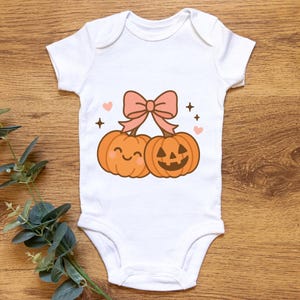 May include: White baby onesie featuring a cartoon design of two orange pumpkins with smiling faces and a pink bow. The design includes pink hearts and black star accents, perfect for Halloween.