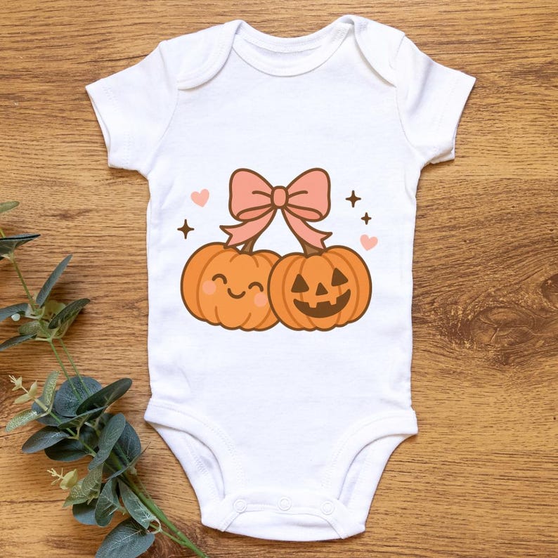 May include: White baby onesie featuring a cartoon design of two orange pumpkins with smiling faces and a pink bow. The design includes pink hearts and black star accents, perfect for Halloween.