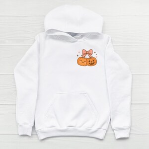 May include: White hooded sweatshirt featuring a front pocket and a drawstring hood. The design on the chest includes two smiling orange pumpkins, a pink bow, hearts, and stars. A cozy and casual garment.