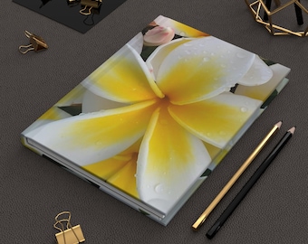 Può includere: Un diario con copertina rigida con un'immagine ravvicinata di un fiore di plumeria bianco e giallo con goccioline d'acqua. Matite dorate e nere, insieme a fermagli dorati, sono disposte attorno al diario su una superficie grigio scuro.