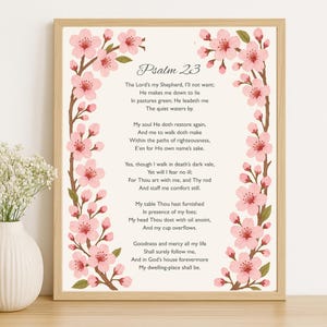 May include: A framed print featuring the text of Psalm 23, surrounded by a floral border of pink cherry blossoms on brown branches. The artwork is set against a cream-colored background, with a light brown wooden frame.