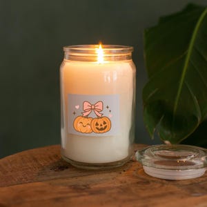 May include: A lit, cream-colored candle in a glass jar with a decorative label featuring two cartoon pumpkins and a pink bow. The candle sits on a wooden surface, with the clear glass lid beside it. A green plant is in the background.