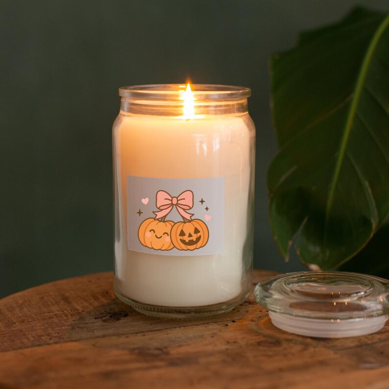 May include: A lit, cream-colored candle in a glass jar with a decorative label featuring two cartoon pumpkins and a pink bow. The candle sits on a wooden surface, with the clear glass lid beside it. A green plant is in the background.
