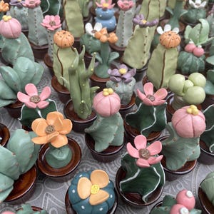 May include: A display of ceramic cactus sculptures in various shapes and colours. The cacti feature green, blue, and pink hues, with some adorned with orange and pink flower accents. Each cactus is potted in a small, brown ceramic pot.