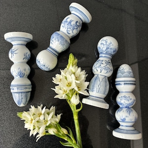 May include: Five white and blue ceramic bottle stoppers with floral patterns. The stoppers are arranged around a cluster of white flowers with green stems. The stoppers have a bulbous shape and are decorated with blue floral patterns.