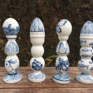 May include: Four white ceramic candle holders with blue hand-painted designs. Each holder has a unique pattern, including waves, leaves, flowers, and fish. The holders are displayed on a wooden surface.
