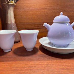 Könnte beinhalten: Eine Lavendel-Teekanne mit Deckel und passendem Untersetzer sowie zwei weiße Teetassen. Die Teekanne und die Tassen stehen auf einer Holzoberfläche.