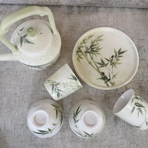 Puede incluir: Una tetera blanca con un diseño de bambú verde y un juego a juego de tazas de té y un platillo. La tetera tiene un asa y un pico. Las tazas de té y el platillo tienen un diseño de bambú verde.