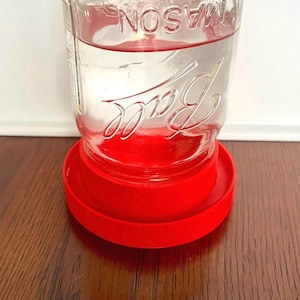 Peut inclure: Un bocal Mason Ball en verre transparent avec le mot "MASON" en haut et "Ball" en script sur le devant. Le bocal repose sur une base en plastique rouge vif. Le bocal est rempli de liquide clair.