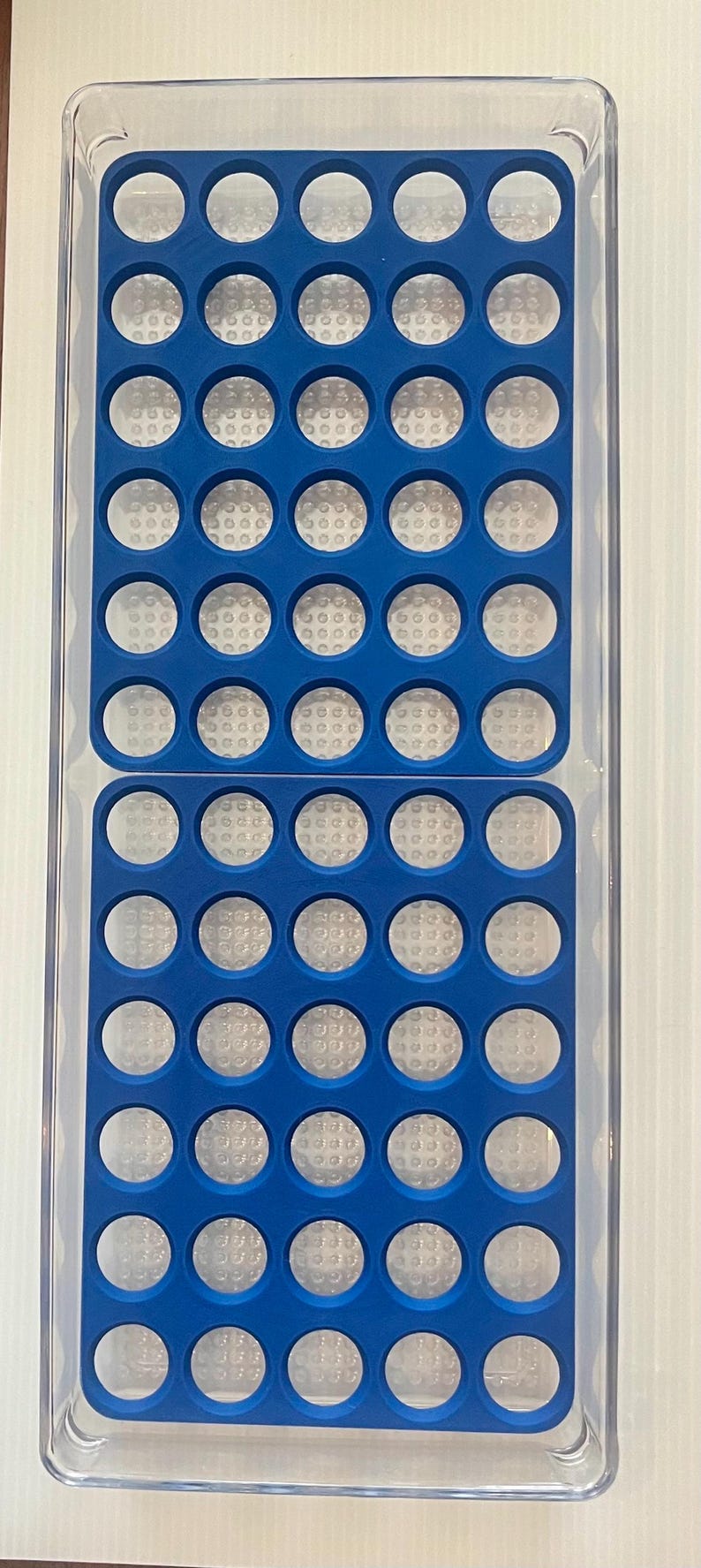 May include: A clear plastic storage container with a blue silicone insert. The insert has 50 round compartments.