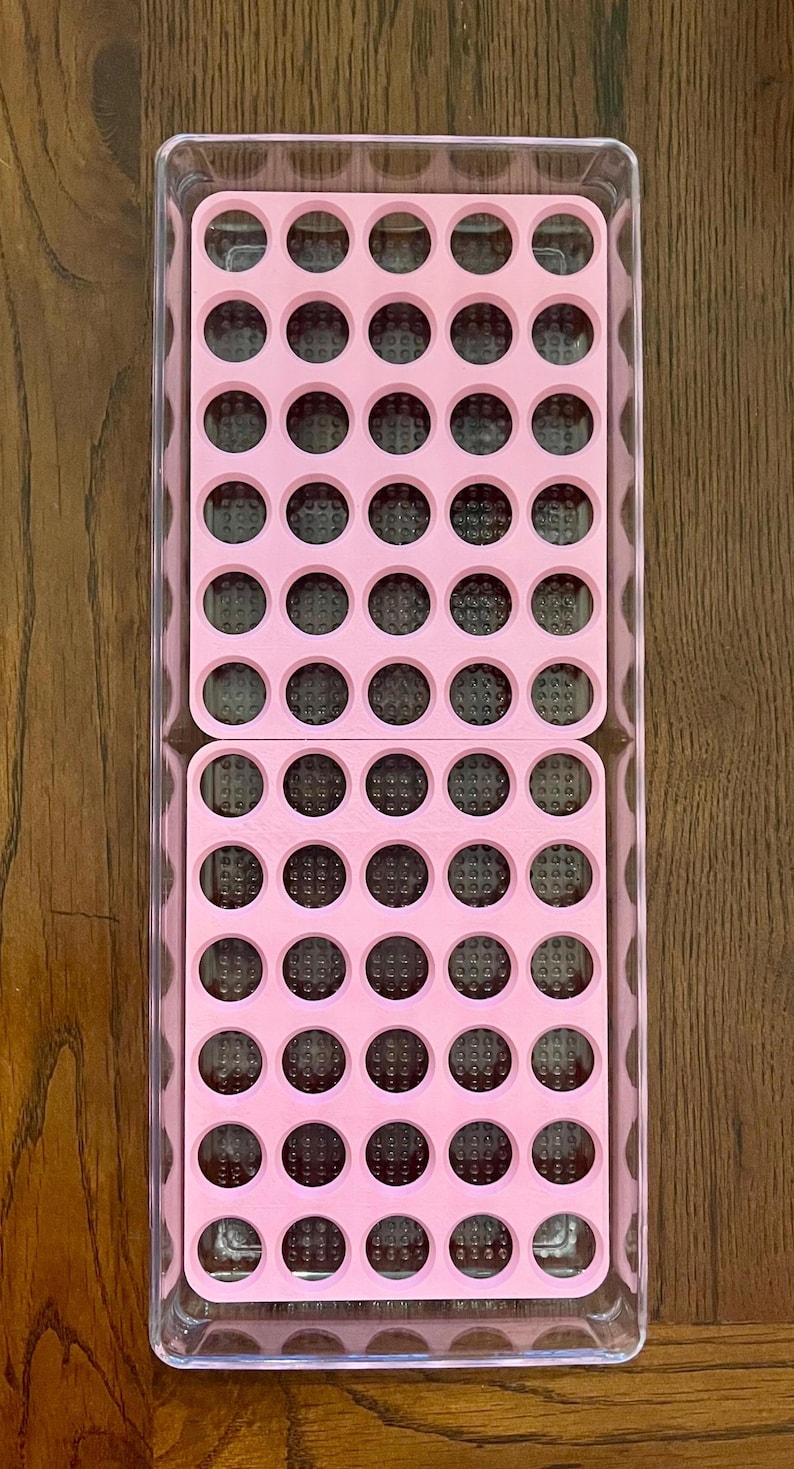 May include: A clear plastic storage container with a pink silicone insert. The insert has 48 round compartments.