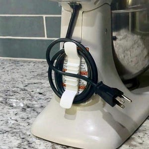 May include: Close-up of a white stand mixer with a black power cord wrapped around a white cord holder. The cord is plugged into an electrical outlet. The mixer is white and has a stainless steel bowl.