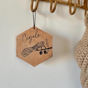 Puede incluir: Un letrero hexagonal de madera con una cuerda negra colgando de la parte superior. El letrero tiene un fondo marrón y una ilustración negra de una cigarra en una rama con dos aceitunas negras. El letrero dice "Cigale" en escritura cursiva negra.