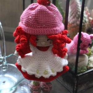 May include: A crocheted doll with red hair, a pink hat with a strawberry on top, and a white dress with red trim. The doll is standing in front of a miniature garden scene.