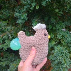 May include: A brown crocheted chicken toy with a white crocheted hat and a blue and green egg. The toy is being held in a hand.