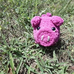 May include: A pink crocheted pig toy with black eyes and a white snout. The toy is sitting on a bed of green grass.