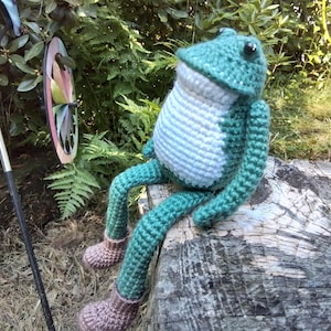 May include: A green crocheted frog with white accents sits on a tree stump. The frog has large, black eyes and is wearing brown crocheted boots.