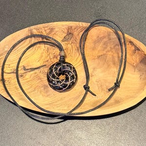 Könnte beinhalten: Ein schwarzer, kreisförmiger Anhänger mit einem geometrischen Muster aus goldfarbenem Draht. Der Anhänger hängt an einer dünnen schwarzen Kordelkette mit einer kleinen metallischen Perle direkt über dem kreisförmigen Element.