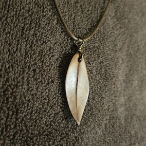 May include: A wooden leaf pendant necklace. The pendant is a light brown, elongated oval shape with a central vein detail. It hangs from a thin, tan cord with a brass-colored clasp. The background is a textured, brown fabric.