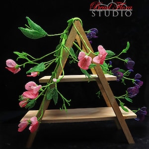 Könnte beinhalten: Eine Perlenblumen-Arrangement mit rosa und violetten Duftenden Erbsenblüten mit grünen Blättern. Die Blumen werden auf einem dreistufigen Holzständer präsentiert. Der Text "Bead Flora STUDIO" ist in der oberen rechten Ecke des Bildes sichtbar.