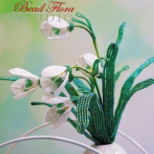 Könnte beinhalten: Ein Strauß weißer Perlen-Schneeglockchen mit grünen Perlen-Stielen in einer weißen Keramikvase. Die Blumen sind in einer Vase mit einem weißen Metallständer arrangiert. Der Text "Bead Flora Studio" ist im Hintergrund sichtbar.