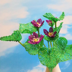 May include: A bouquet of beaded flowers with green leaves in a white vase. The flowers are purple and pink with yellow centers. The background is a light blue sky with white clouds. The text "Bead Flora Studio" is at the bottom of the image.