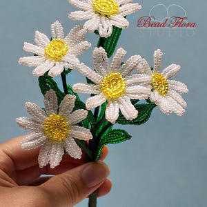 May include: A bouquet of five white beaded daisies with yellow centers and green beaded leaves. The daisies are arranged in a hand-held bouquet.