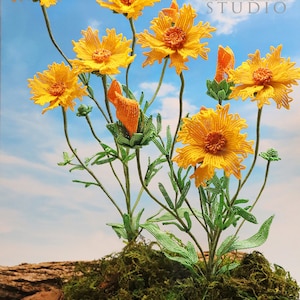 Puede incluir: Un ramo de flores amarillas de cuentas con tallos y hojas verdes sobre un fondo de cielo azul. Las flores están dispuestas en un jarrón sobre un lecho de musgo. El texto "Bead Flora Studio" es visible en la esquina superior derecha de la imagen.