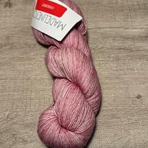 May include: A skein of pink and white variegated yarn with a label that reads "MADELINETOSH". The yarn is tightly wound and appears soft. The label is red and white. The yarn is on a wooden surface.