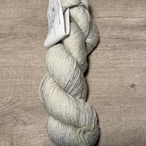 May include: A skein of light gray yarn with a subtle sheen, tightly wound and ready for knitting or crocheting. A white tag is attached to the top of the yarn. The yarn is displayed on a light brown wooden surface.