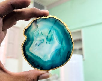 May include: A round, polished agate slice with a gold-colored edge. The stone features concentric bands of teal and white, creating a natural, swirling pattern. The agate is held up by a hand, showcasing its unique, organic design.