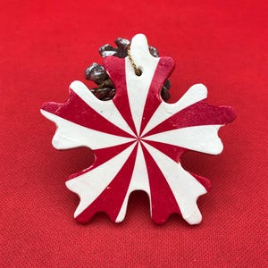 May include: A snowflake-shaped Christmas ornament with red and white stripes radiating from the center. The ornament is attached to a small pine cone and has a gold string for hanging. The background is a solid red.