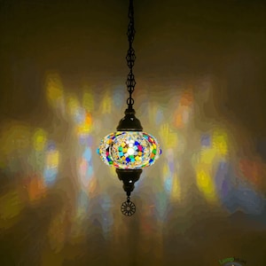 Op de afbeelding: Een kleurrijke moza&iuml;ek glazen hanglamp met een ronde, bolvormige kap. De lamp hangt aan een ketting en heeft een metalen voet.