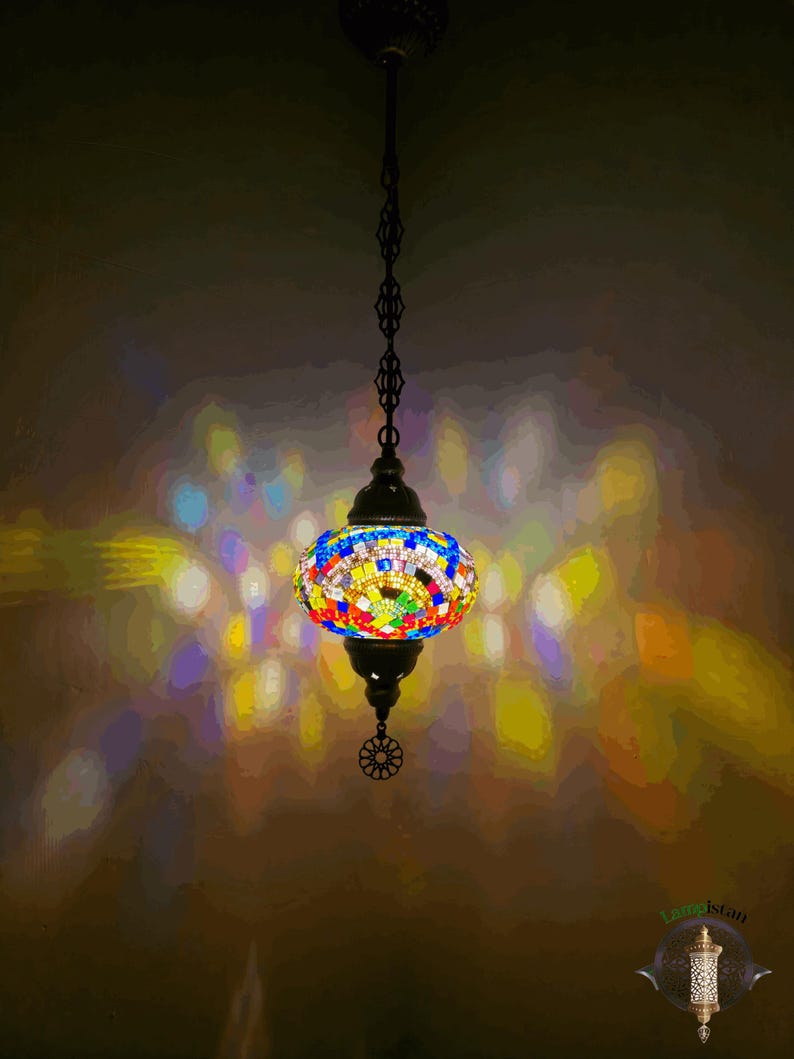 Op de afbeelding: Een kleurrijke moza&iuml;ek glazen hanglamp met een zwarte metalen ketting en een decoratief metalen eindstuk. Het licht werpt een kleurrijk patroon op het plafond.
