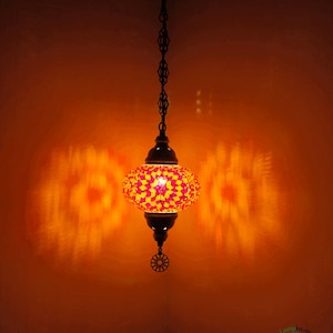 Op de afbeelding: Een hangende moza&iuml;ek glazen lamp met een geometrisch patroon in roze en geel. De lamp is aan en werpt een warme gloed op de muur.