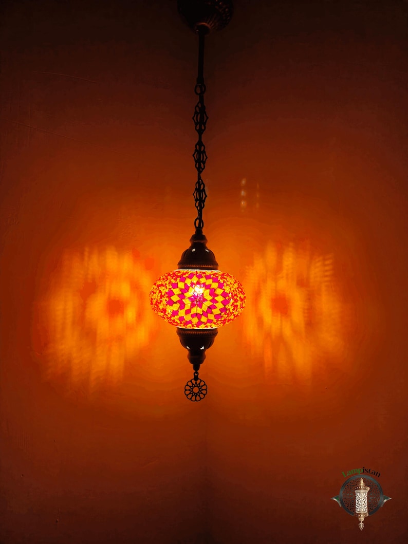 Op de afbeelding: Een hangende moza&iuml;ek glazen lamp met een geometrisch patroon in roze en geel. De lamp is aan en werpt een warme gloed op de muur.