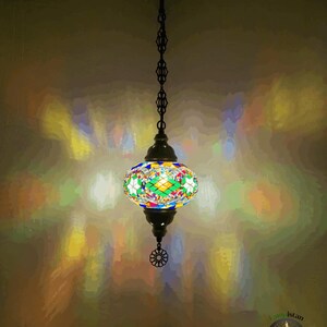 Op de afbeelding: Een kleurrijke moza&iuml;ek glazen hanglamp hangt aan een ketting. Het licht werpt een regenboogpatroon op de muur.