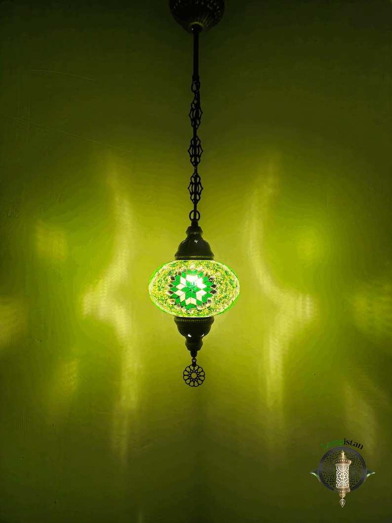 Op de afbeelding: Een groene en witte moza&iuml;ek glazen hanglamp met een zwarte metalen ketting en een decoratief metalen eindstuk. De lamp hangt aan een plafond.