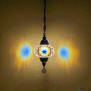 Op de afbeelding: Een kleurrijke moza&iuml;ek glazen hanglamp hangt aan een ketting. De lamp is rond en heeft een stervormig ontwerp in het midden. Het licht is aan en geeft een warm gloed.
