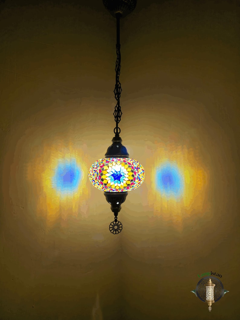 Op de afbeelding: Een kleurrijke moza&iuml;ek glazen hanglamp hangt aan een ketting. De lamp is rond en heeft een stervormig ontwerp in het midden. Het licht is aan en geeft een warm gloed.